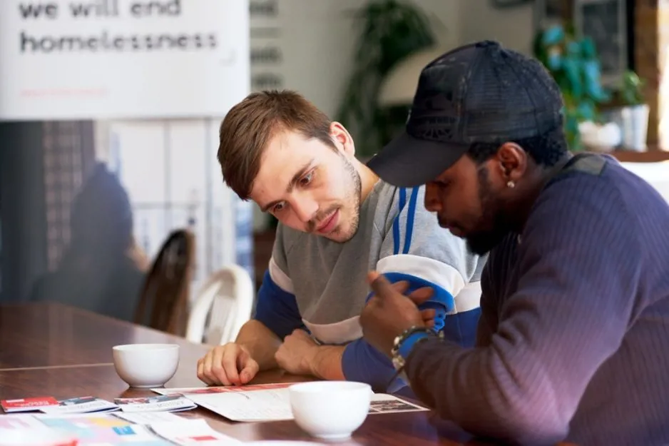 A photo of two men sitting at a table next to each other looking at a leaflet, with coffee cups in the foreground