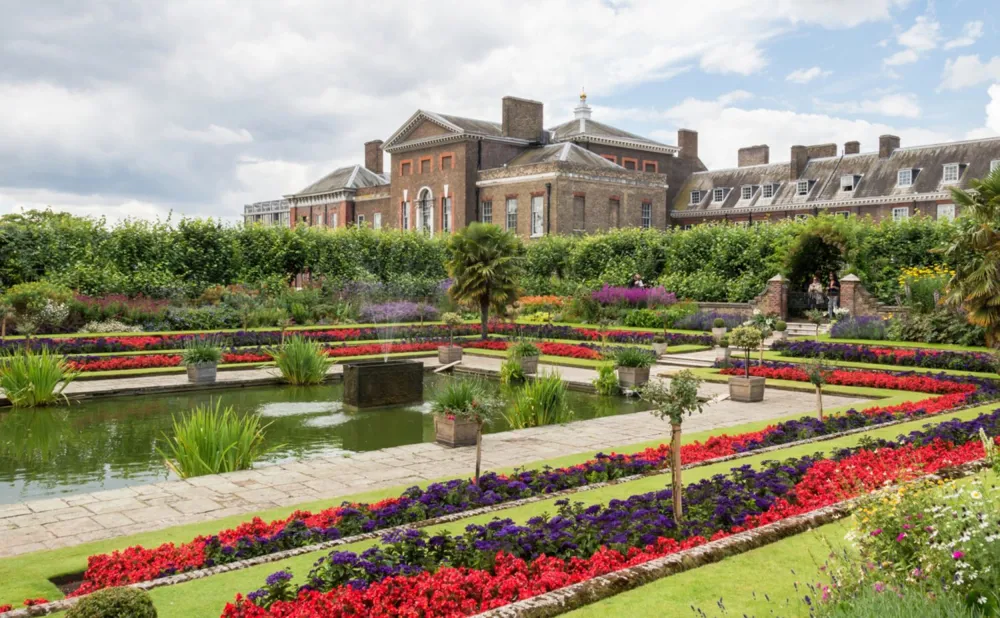 A photo of a fountain in a garden with a large building in the background